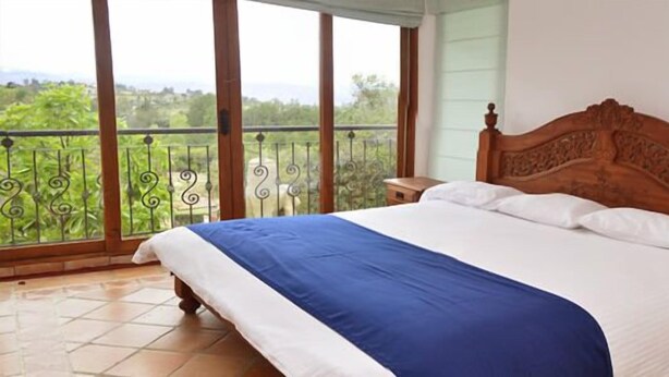 A bedroom with a large bed, a wooden headboard, a nightstand, and a view of greenery through a balcony. - Hotel Campestre Bachue (Villa de Leyva)