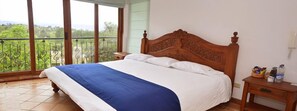 Un dormitorio con una cama grande, cabecera de madera, mesita de noche y vista a la vegetación desde un balcón.