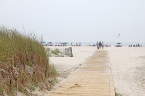 Une promenade en bois mène à une plage de sable où l’on aperçoit des parasols et des gens au loin.