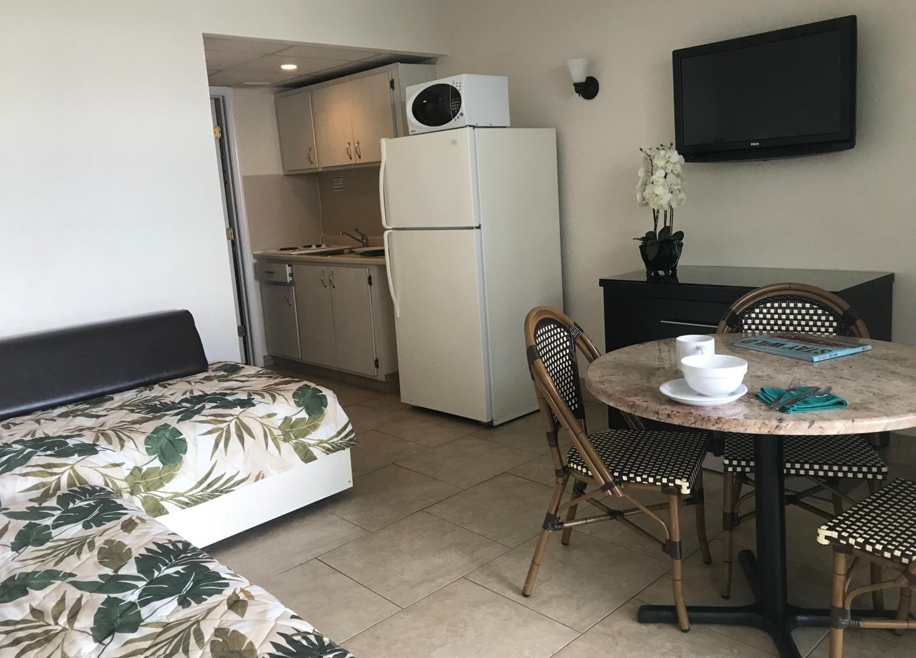 A compact room with a bed, a dining table with chairs, a small kitchen area with a refrigerator, and a flat-screen TV mounted on the wall.