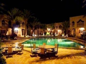 A nighttime view of a hotel pool area with lounge chairs and tables, surrounded by palm trees and illuminated buildings. - Hôtel Erfoud le Riad (Erfoud)