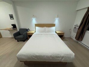 A hotel room with a large bed, bedside tables, a chair, a desk with a coffee maker, and an air conditioning unit.