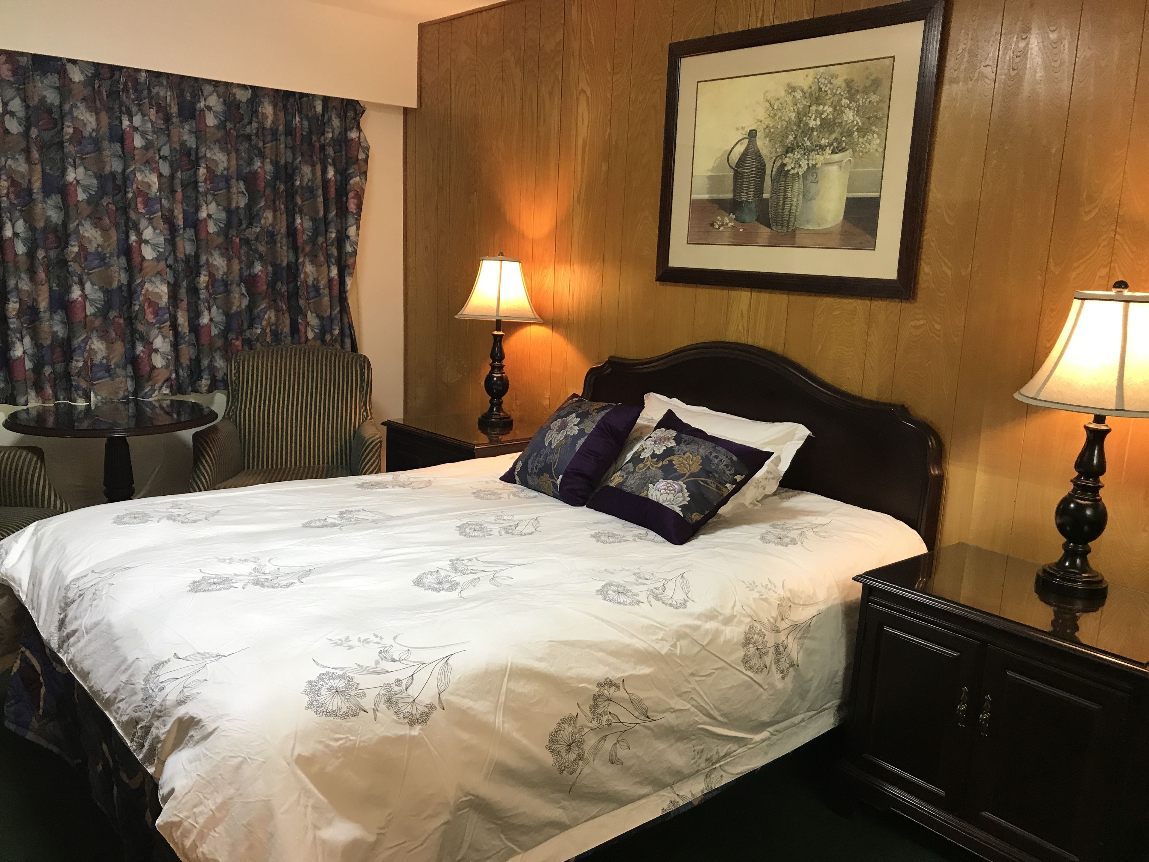 A hotel room with a bed, two bedside lamps, a chair, a small table, and a floral curtain.