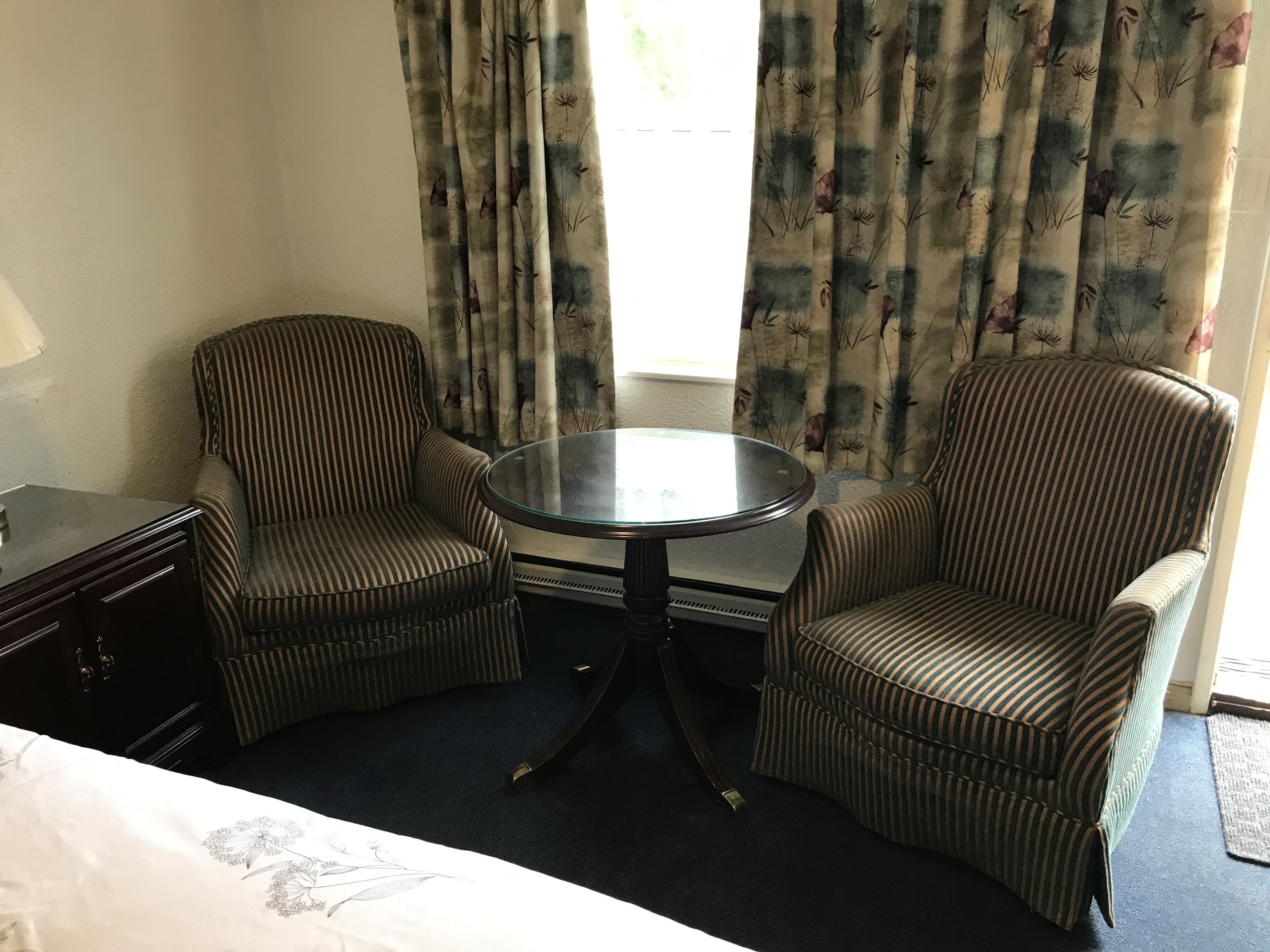 Quarto de hotel com duas poltronas listradas, uma mesa de vidro pequena, uma cama e uma janela com cortinas florais.