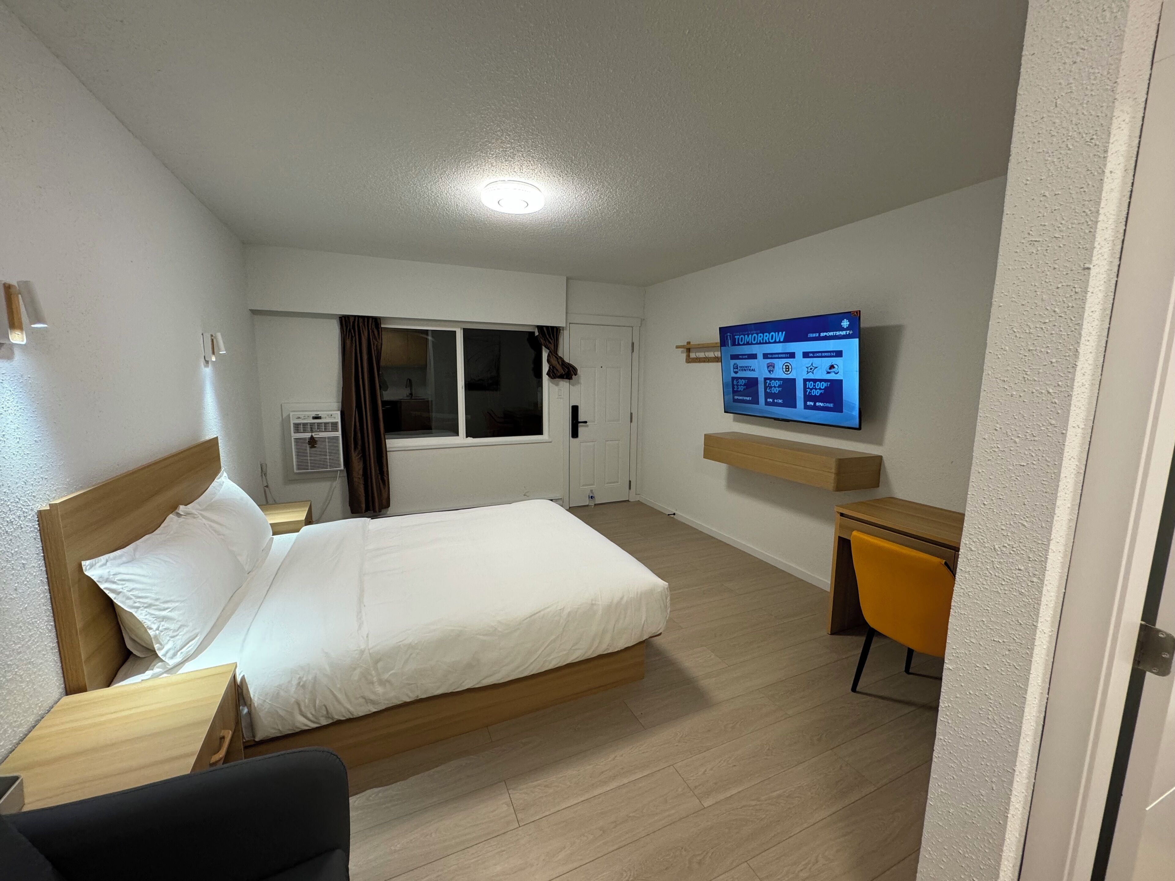 A hotel room with a bed, a desk, a chair, a TV mounted on the wall, and a window with curtains.