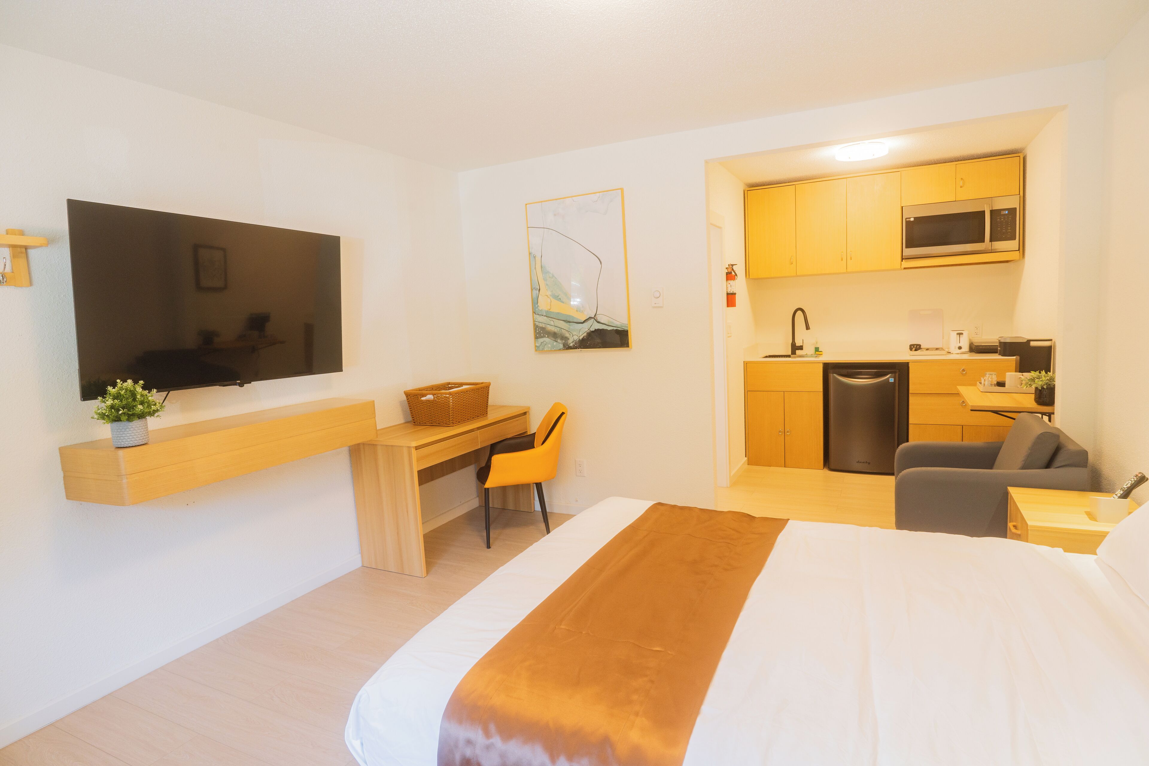 A hotel room with a bed, a desk, a chair, a television, a kitchen area, and a microwave.
