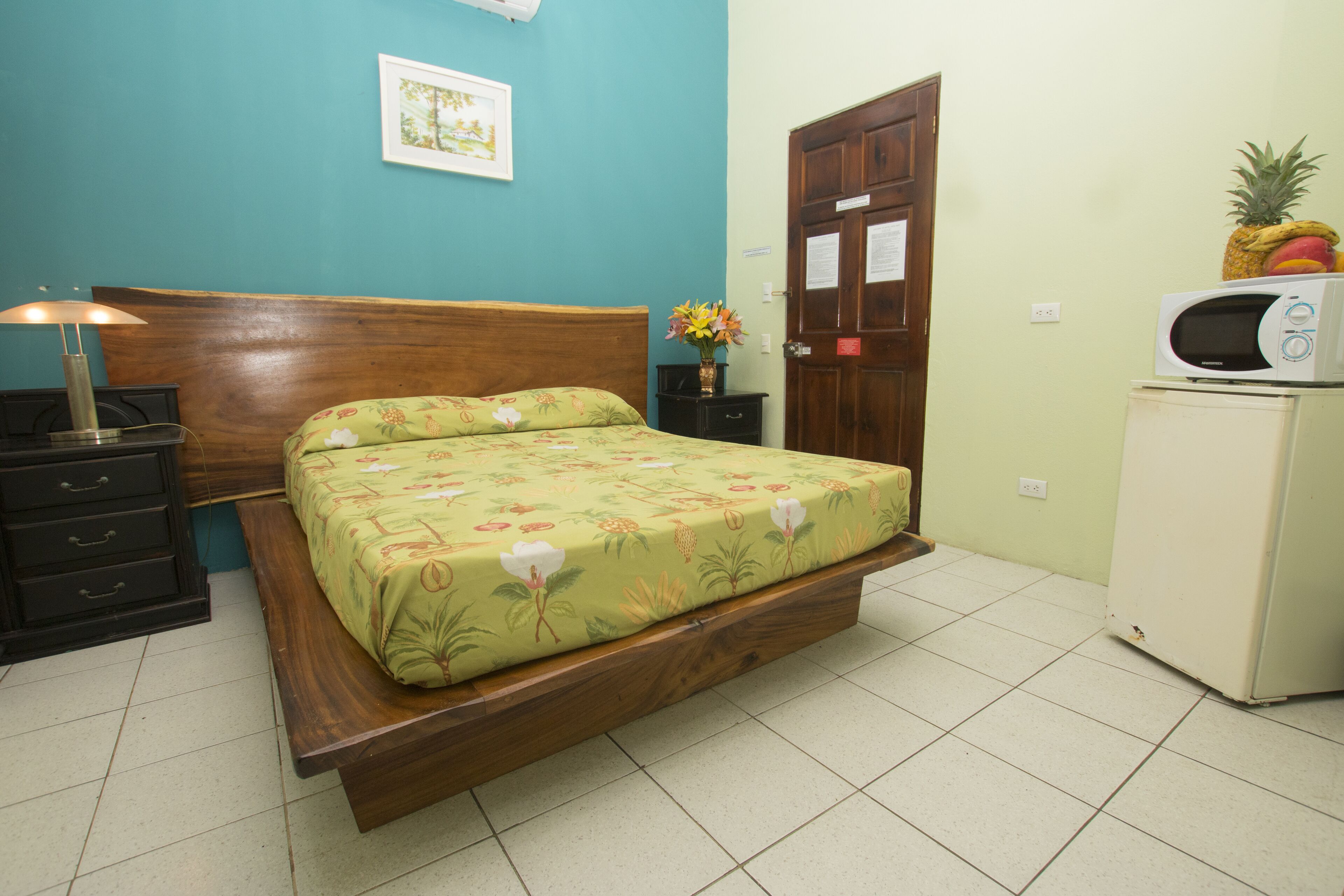 A bedroom with a wooden bed, a nightstand, a lamp, a microwave, and a door.