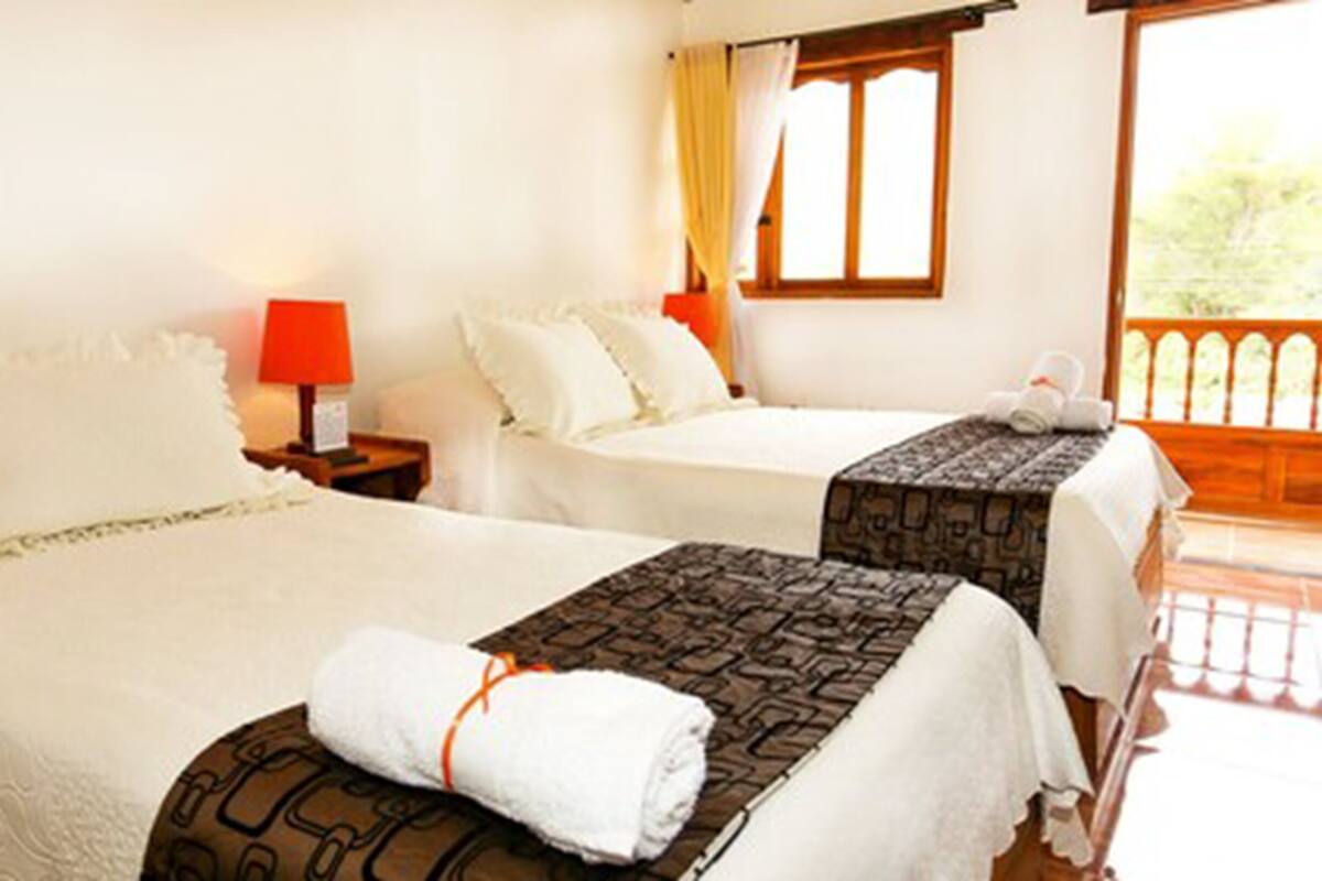 Habitación de hotel con dos camas, cada una con una colcha estampada y una toalla enrollada. Hay una mesita de noche con una lámpara entre las camas.