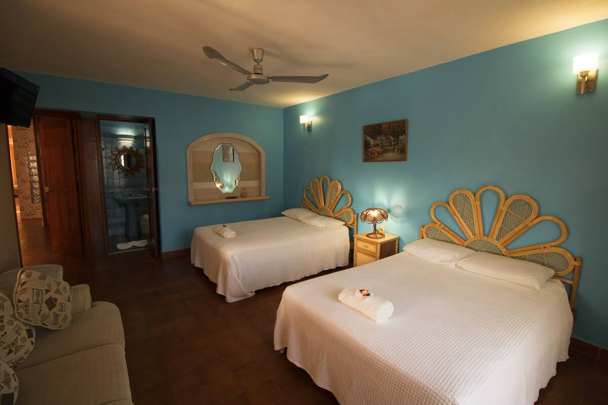 A hotel room with two beds, a ceiling fan, a mirror, a nightstand with a lamp, and a door leading to a bathroom.