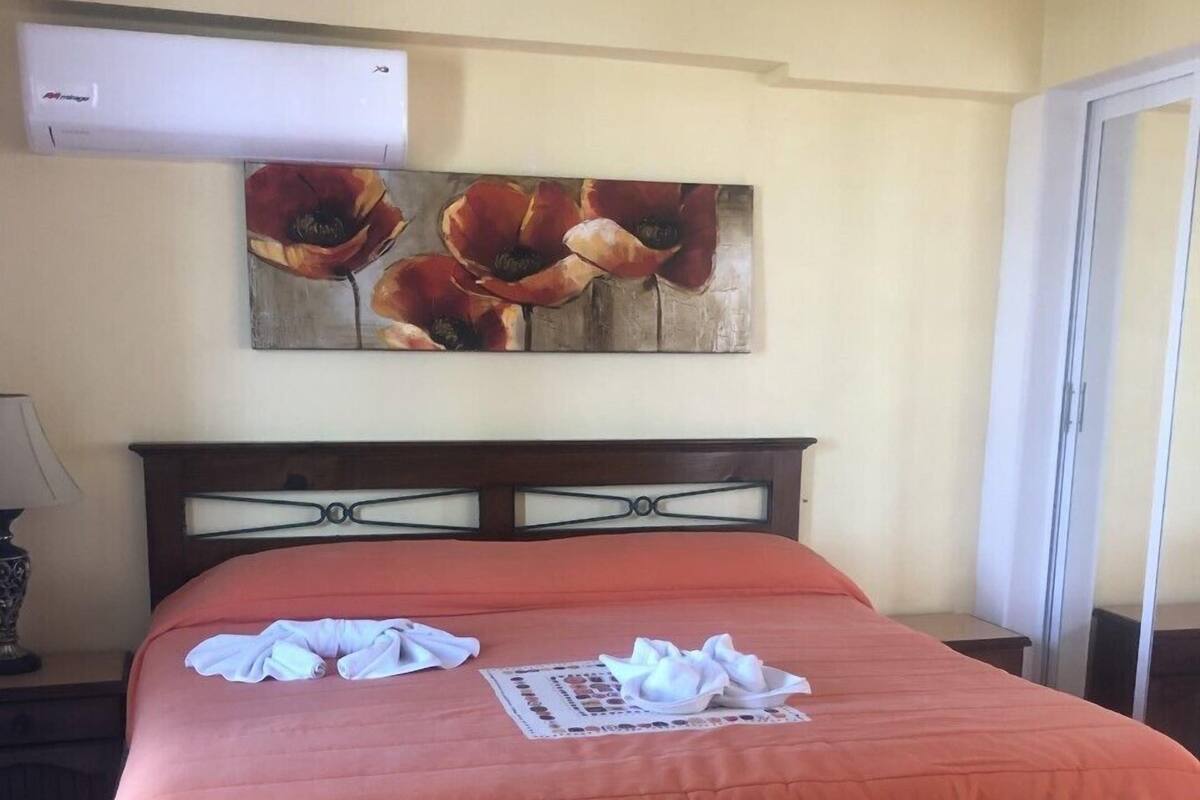 A hotel room with a bed, a wall-mounted air conditioner, and a painting of flowers.