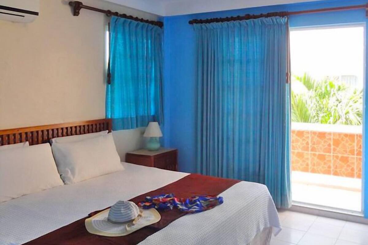 A hotel room with a bed, a nightstand, a lamp, and a view of greenery outside.