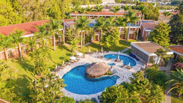 Vista aérea de un complejo turístico con una gran piscina, palmeras y varias edificaciones con tejados rojos.