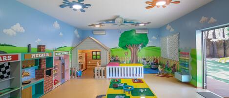 A children's playroom with a play kitchen, tables with toys, and a mural of a landscape.