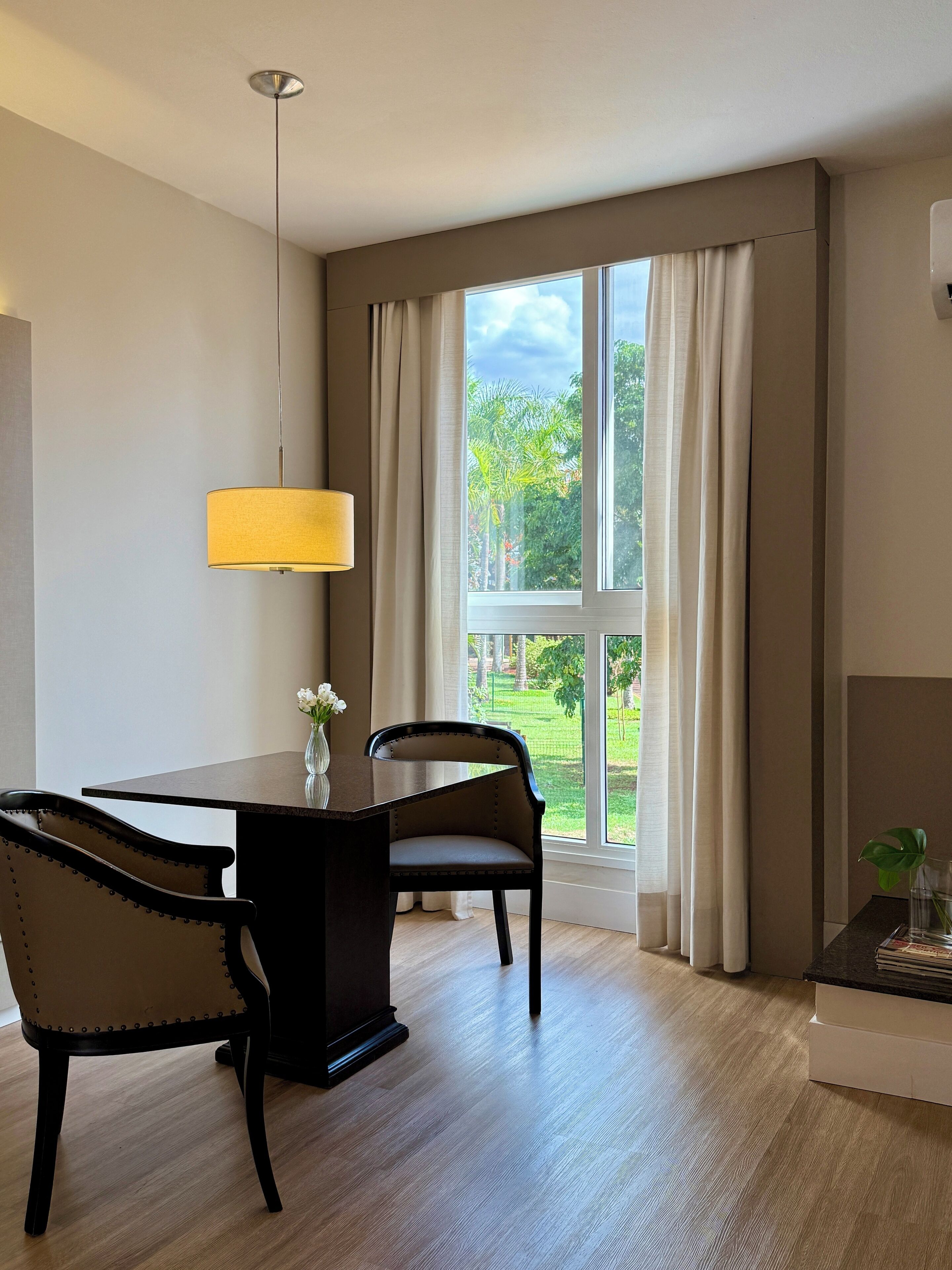 A modern dining area with a wooden table and chairs, a hanging lamp, and a view of a garden through a large window with curtains.