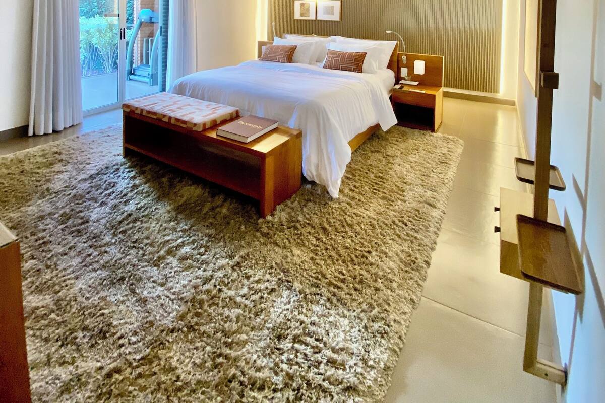 A modern bedroom with a large bed, a wooden bench, a nightstand, and a view of the outdoors.