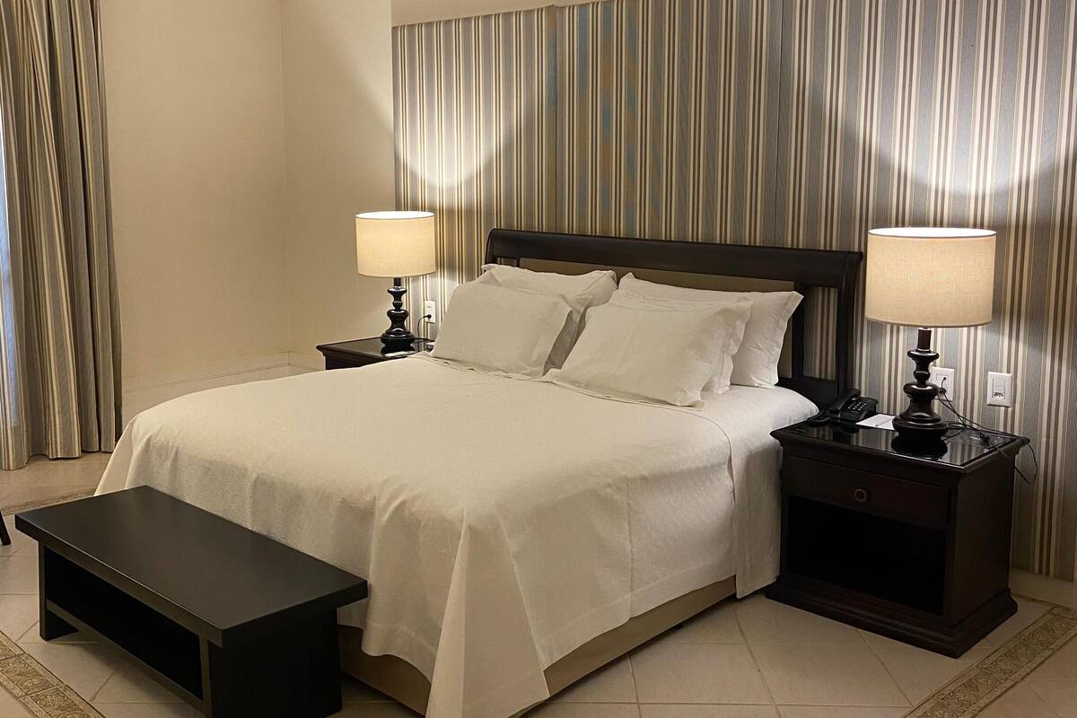 A hotel room with a large bed, two bedside tables with lamps, a bench, and a striped curtain backdrop.