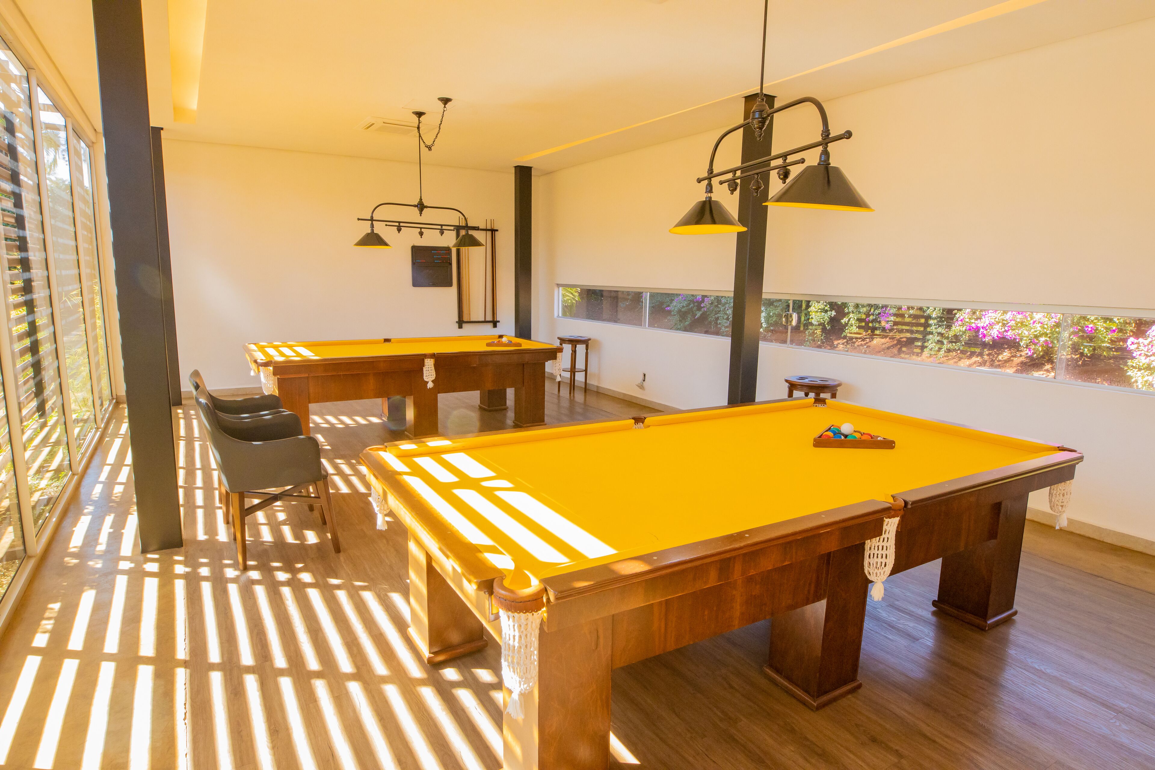 A pool table with a yellow felt surface, a wooden table, and a chair. There are two hanging lamps with black shades.