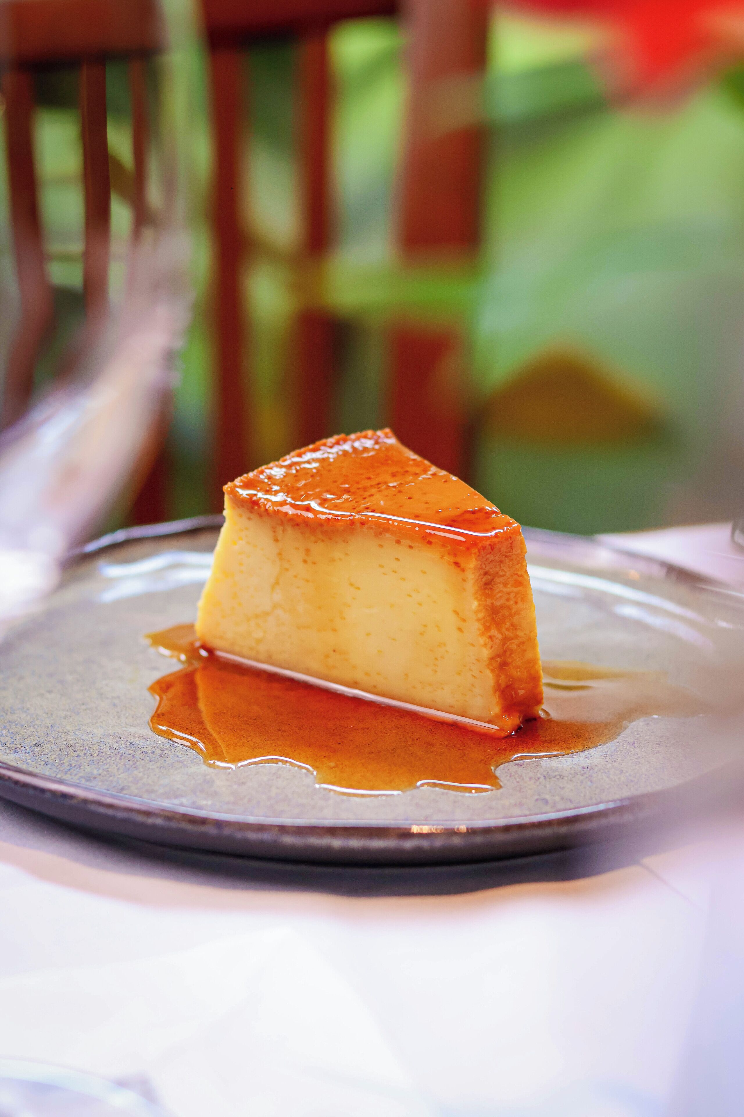 Una porción de flan en un plato con salsa de caramelo, sobre una mesa con un fondo desenfocado.