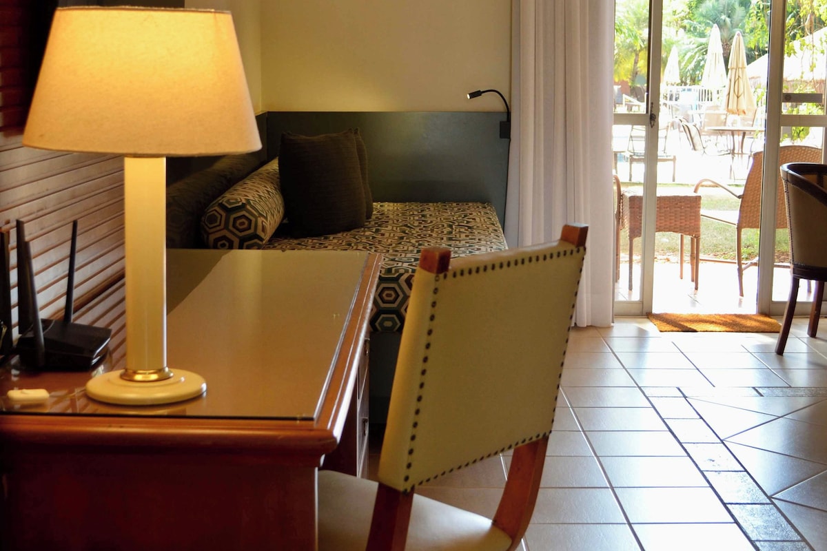 Habitación de hotel con cama, escritorio con lámpara, silla, ventana con cortinas y vista al exterior.