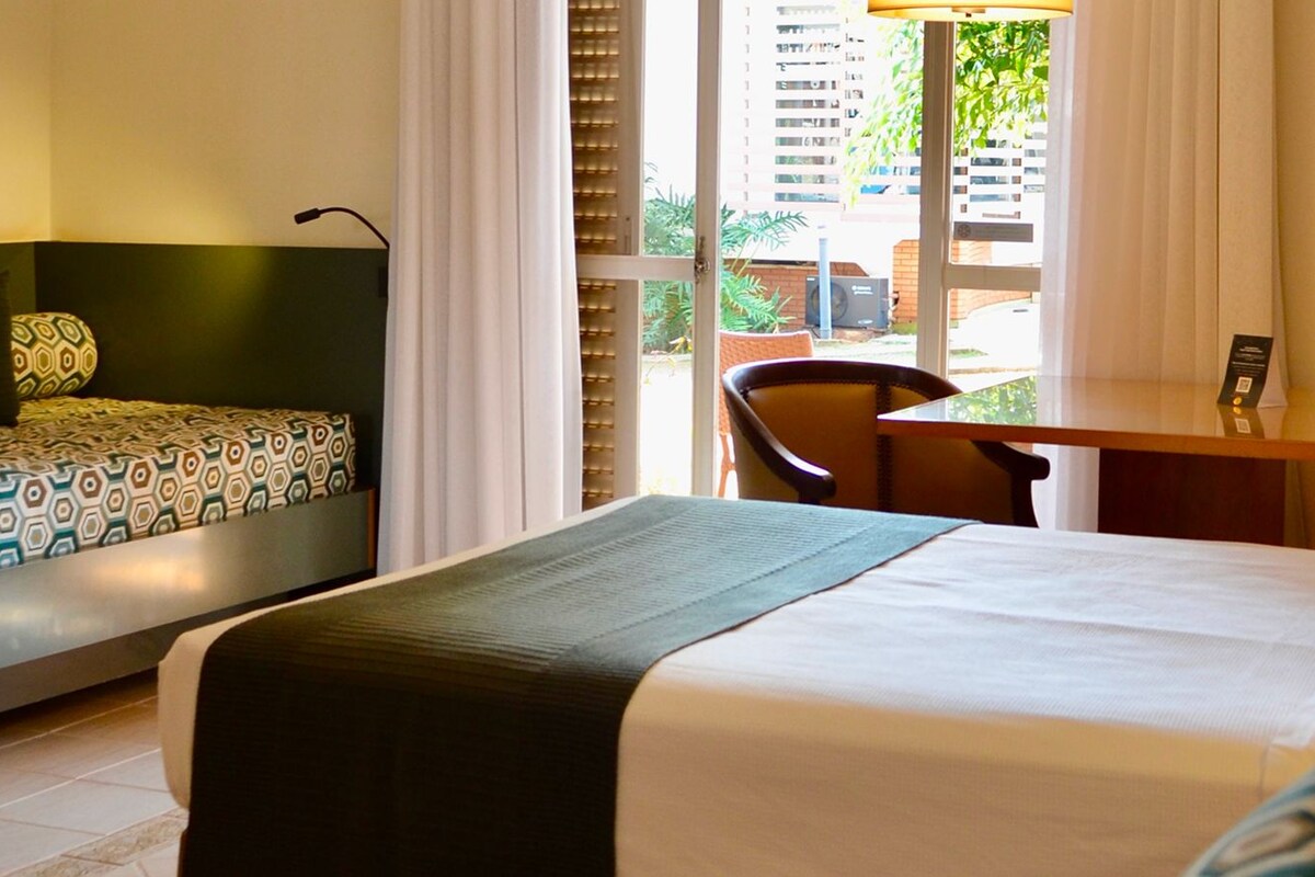 Ein Hotelzimmer mit zwei Betten, einem Schreibtisch und einem Stuhl. Es gibt ein Fenster mit Jalousien und eine Lampe auf dem Schreibtisch.