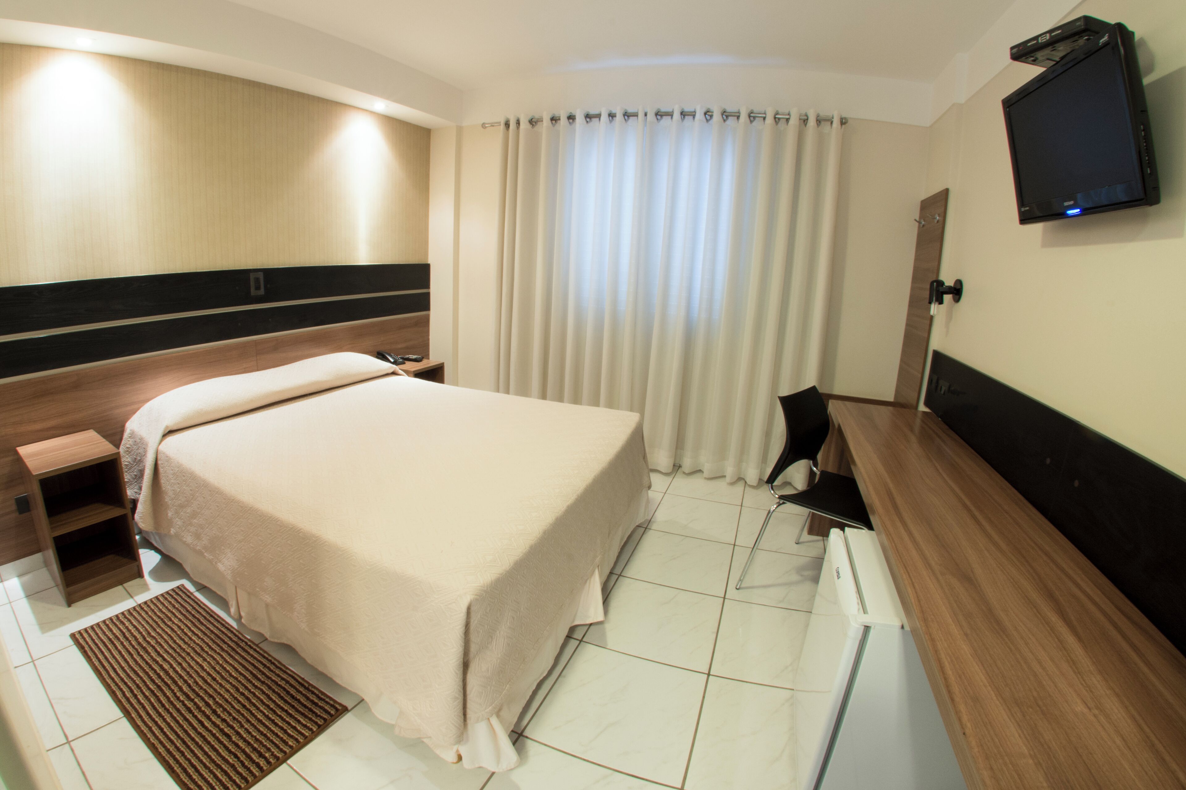 Una habitación de hotel con cama, mesita de noche, televisor montado en la pared, escritorio y ventana con cortinas.