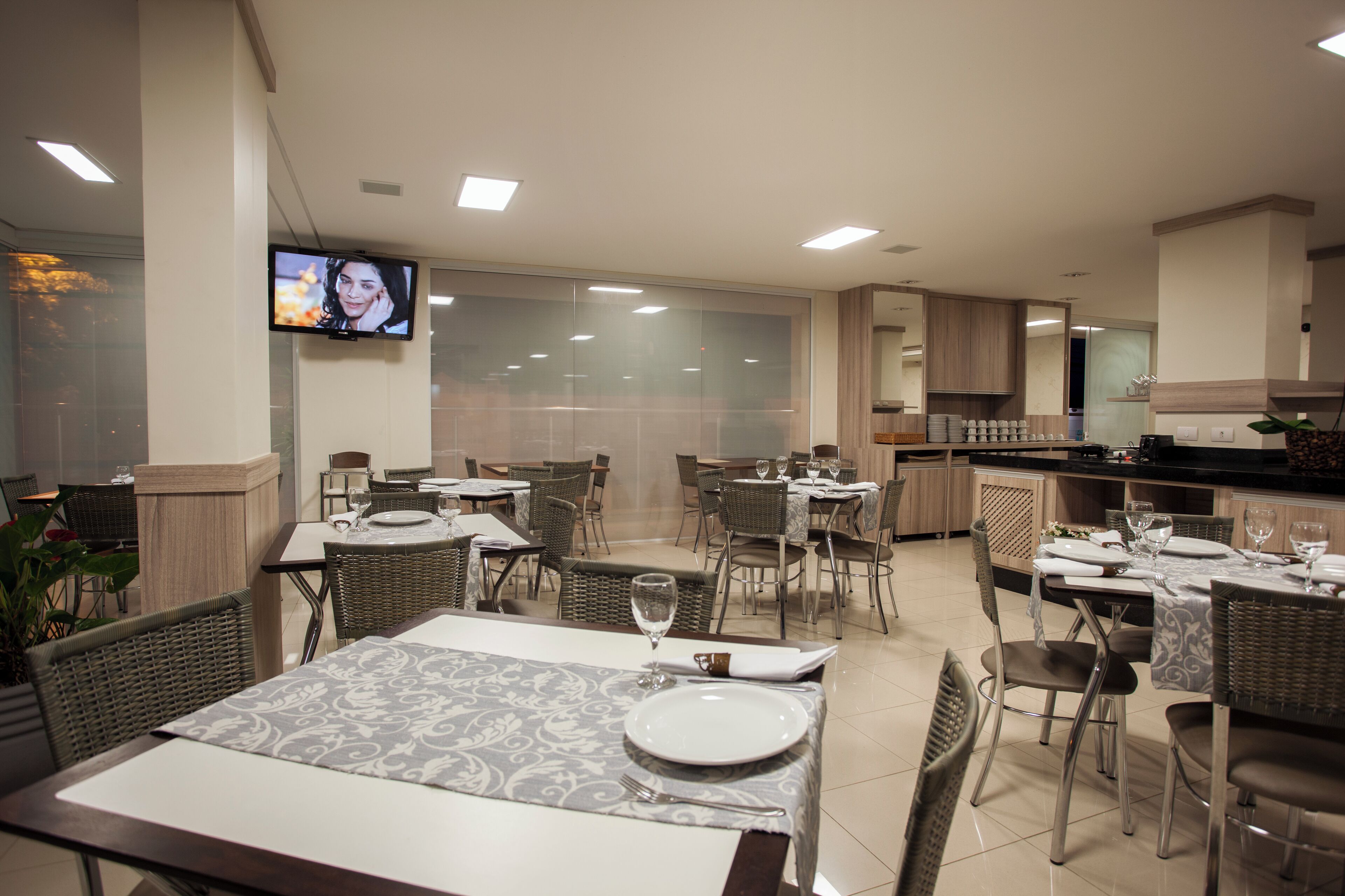 Un restaurante con mesas dispuestas para comer, un televisor colgado en la pared y una zona de cocina al fondo.