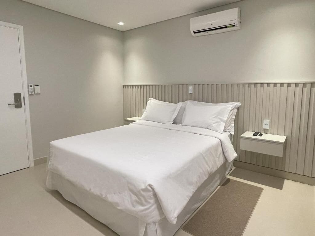 Quarto de hotel moderno com uma cama grande, uma mesinha de cabeceira, um ar condicionado e uma porta com fechadura.