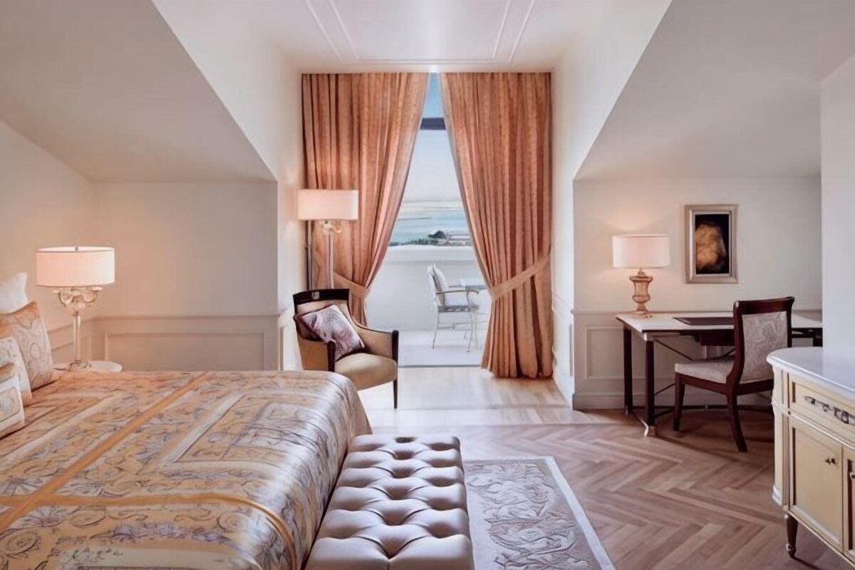A hotel room with a large bed, a desk, a chair, and a view through curtains.