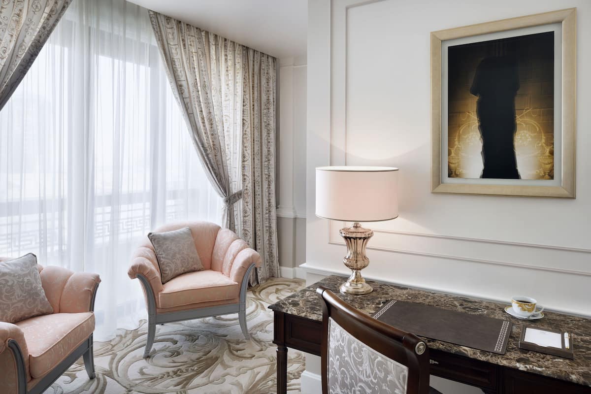 Ein Hotelzimmer mit großem Fenster, zwei bequemen Ohrensesseln, einem Schreibtisch mit Lampe und einem gerahmten Kunstwerk an der Wand.