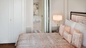 Quarto com cama, mesa de cabeceira, abajur e banheiro visível por uma porta aberta.