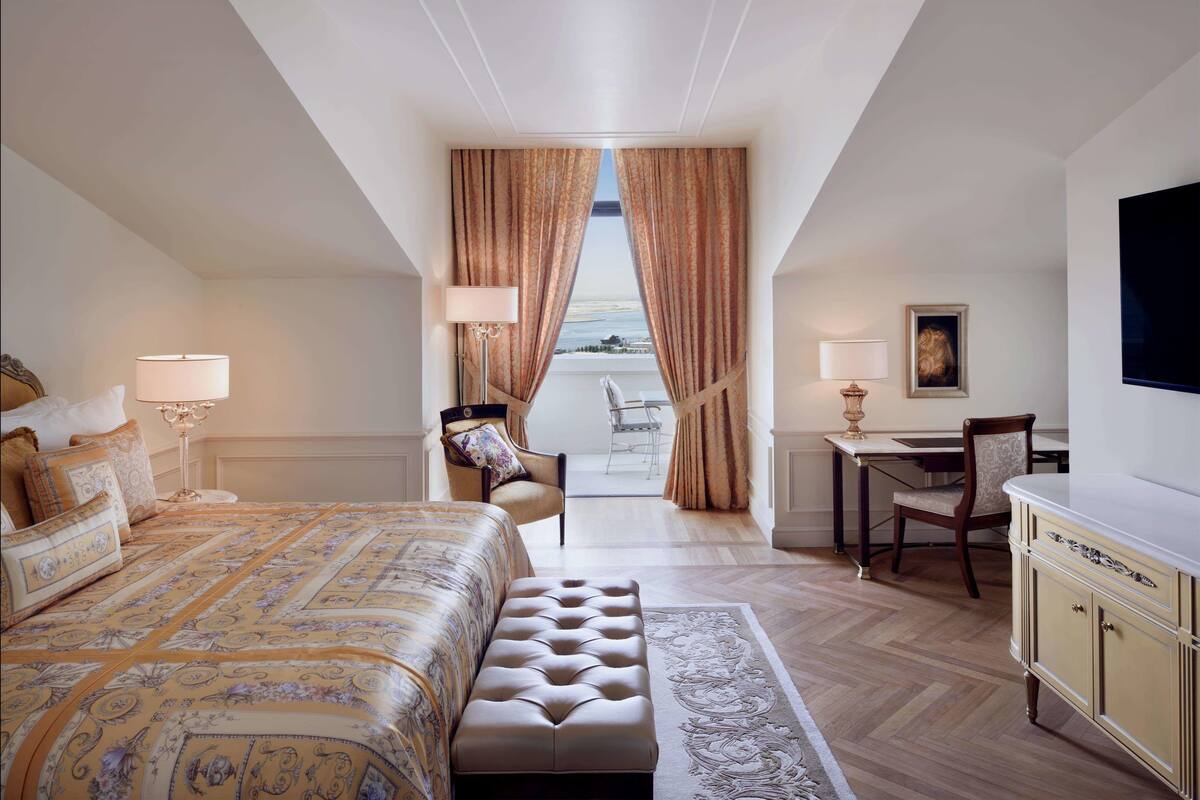 Ein Hotelzimmer mit einem großen Bett, einem Schreibtisch, einem Sessel und Meerblick durch Vorhänge.