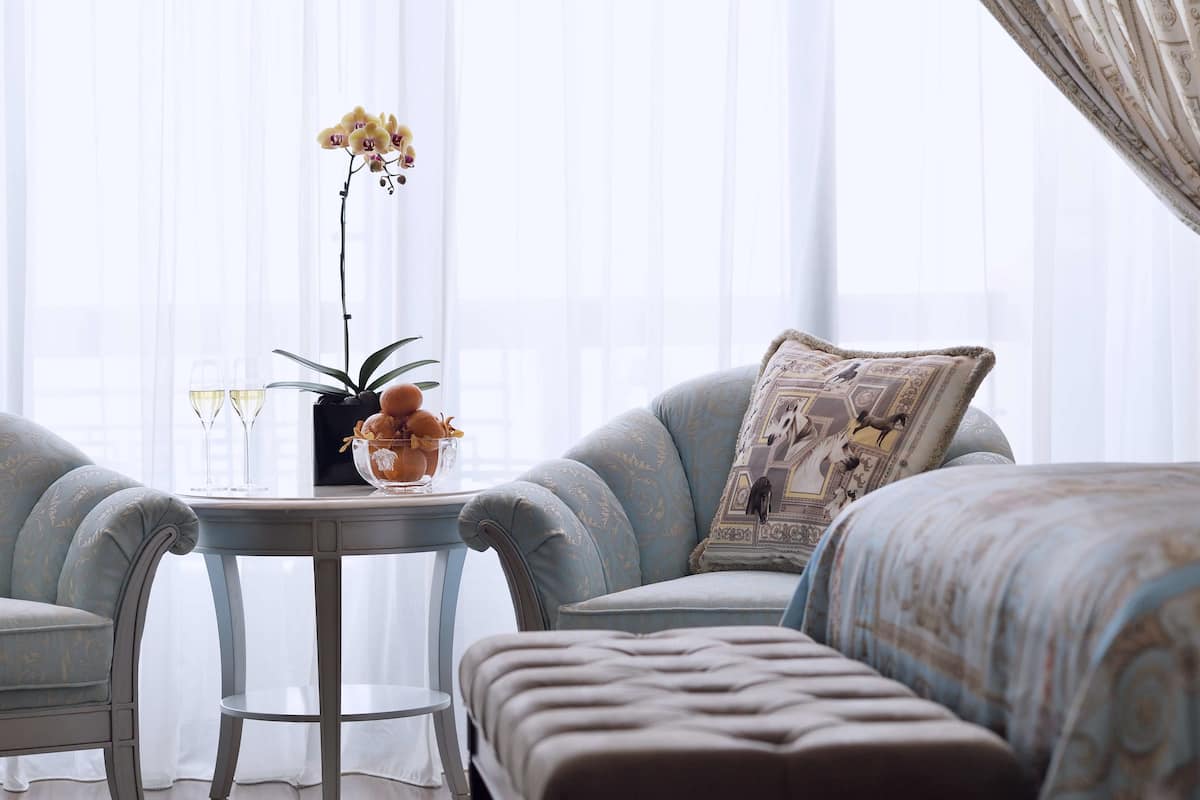 Ein Zimmer mit einem hellblauen Sessel, dazu passendem Hocker, einem kleinen runden Tisch mit Blumenvase und einem Zierkissen.