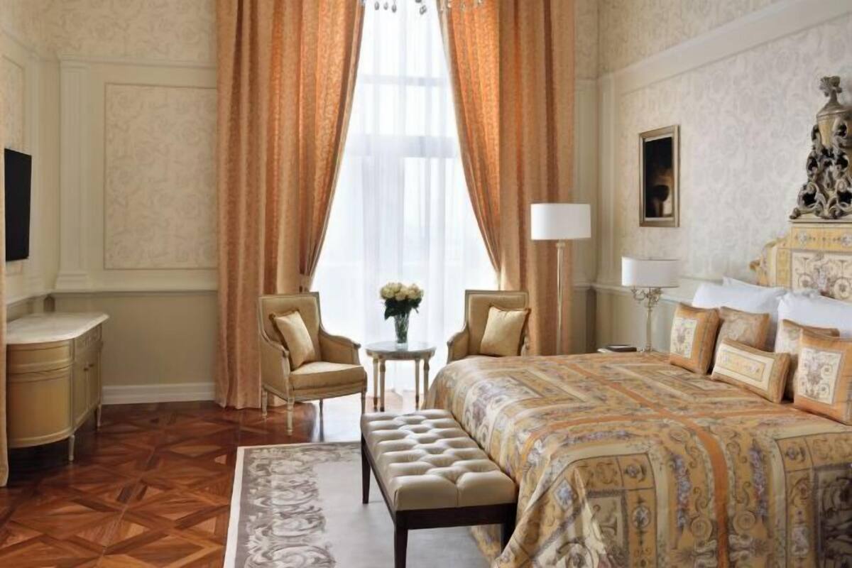 Ein Hotelzimmer mit einem großen Bett, zwei Sesseln, einem kleinen Tisch mit Blumenvase und einer dekorativen Lampe.
