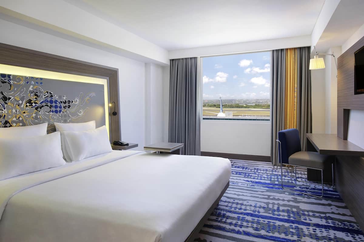 Habitación de hotel con una cama grande, un escritorio, una silla y vista al aeropuerto.