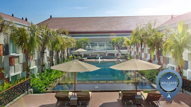 A pool area with lounge chairs and umbrellas, surrounded by palm trees and a modern building with a tiled roof. - Novotel Bali Ngurah Rai Airport (Tuban)