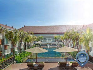 A pool area with lounge chairs and umbrellas, surrounded by palm trees and a modern building with a tiled roof. - Novotel Bali Ngurah Rai Airport (Tuban)
