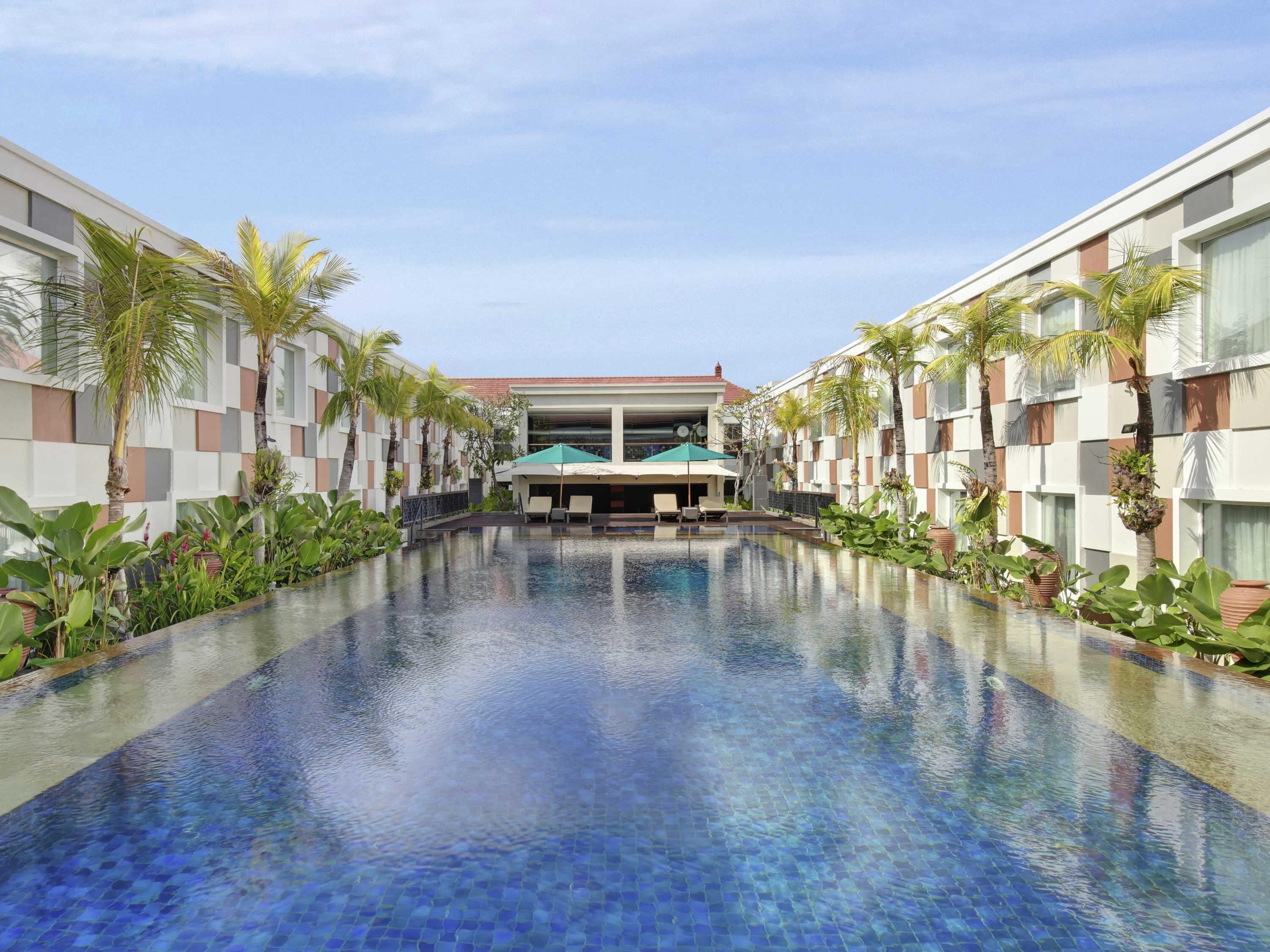 A swimming pool with a tiled floor, surrounded by palm trees and a building with a checkered facade.