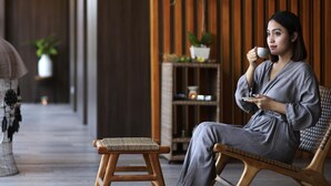 A person sitting on a wicker chair in a room with wooden flooring, a small stool, and a shelf with plants. - Novotel Bali Ngurah Rai Airport (Tuban)