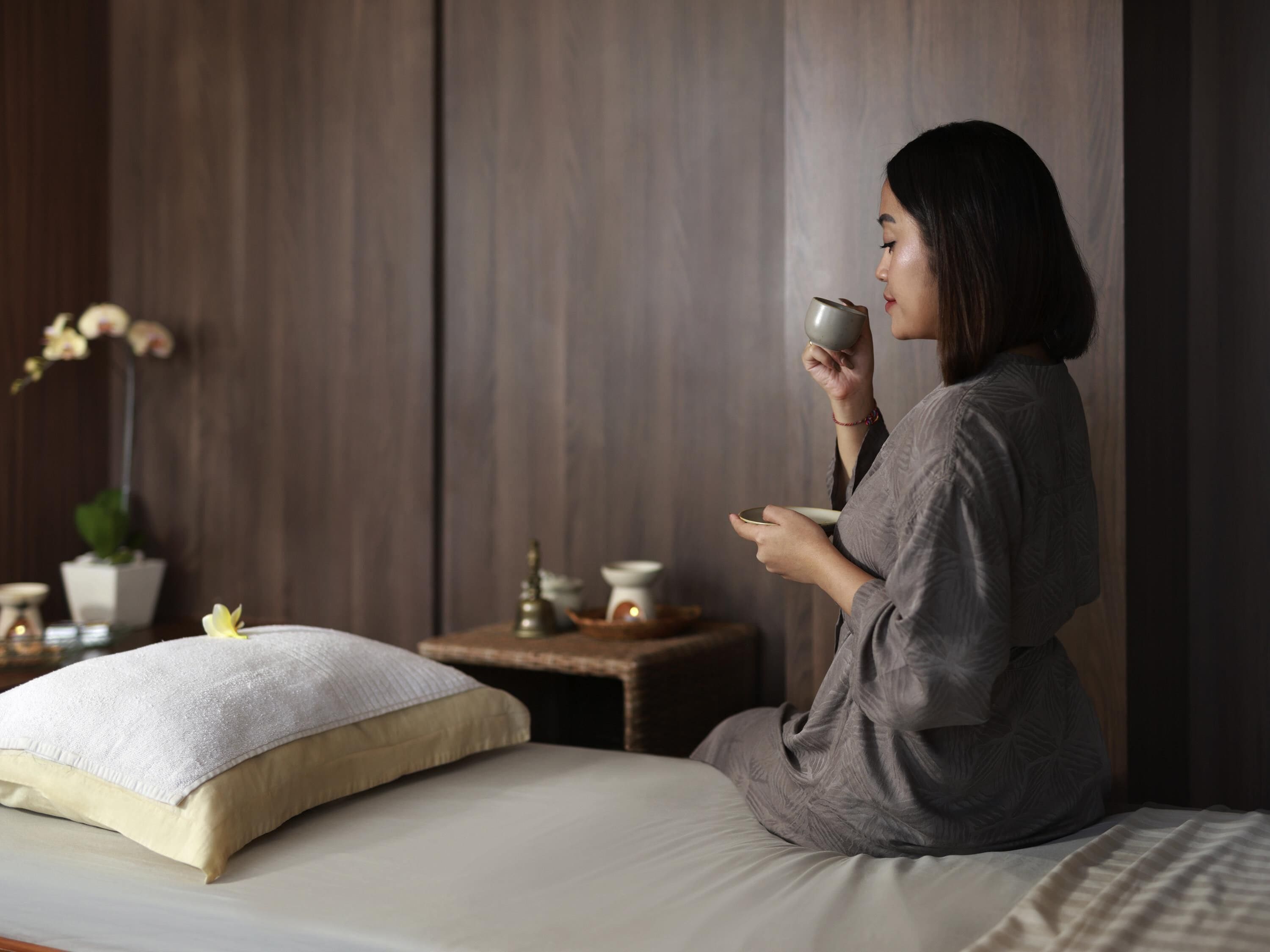 Una persona sentada en una cama en una habitación con pared de madera, una mesita con lámpara y una planta en maceta.