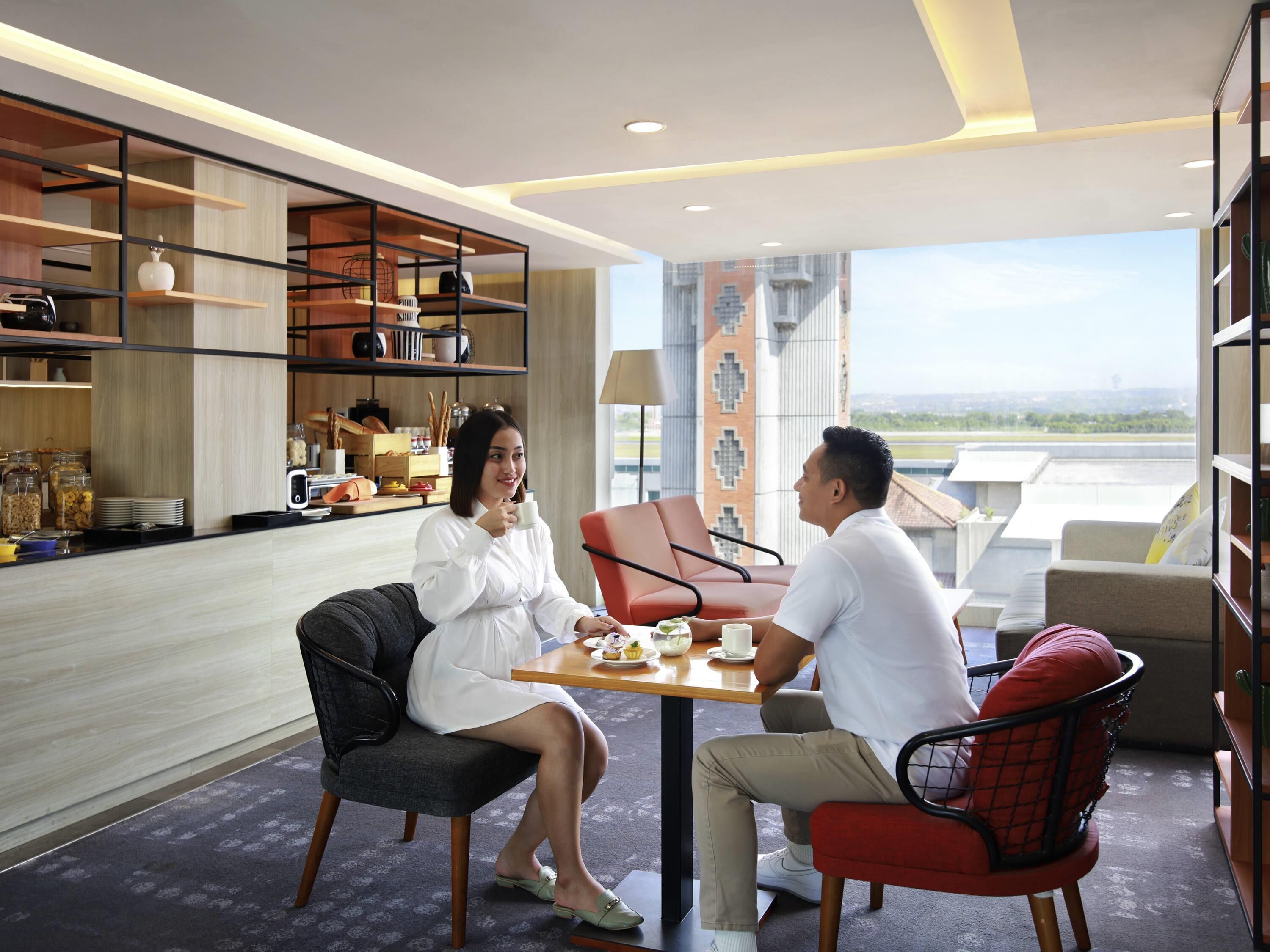 A modern hotel room with a dining area, a large window with a city view, and a bookshelf with various items.