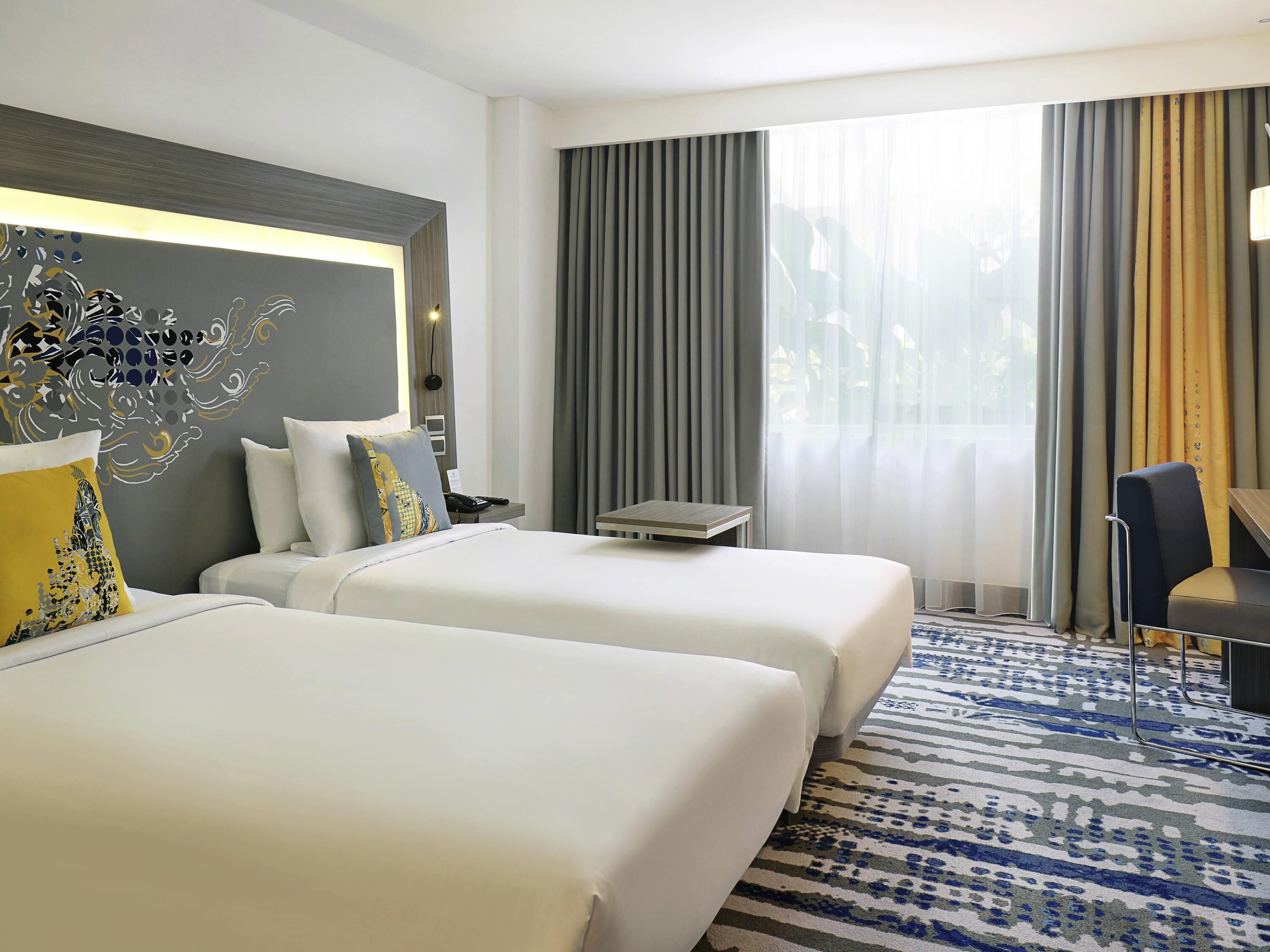 A hotel room with two beds, a desk, a chair, a patterned rug, and a large window with curtains.