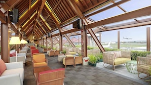 A covered outdoor seating area with various chairs and tables, a wooden ceiling structure, and a view of an airport in the background. - Novotel Bali Ngurah Rai Airport (Tuban)