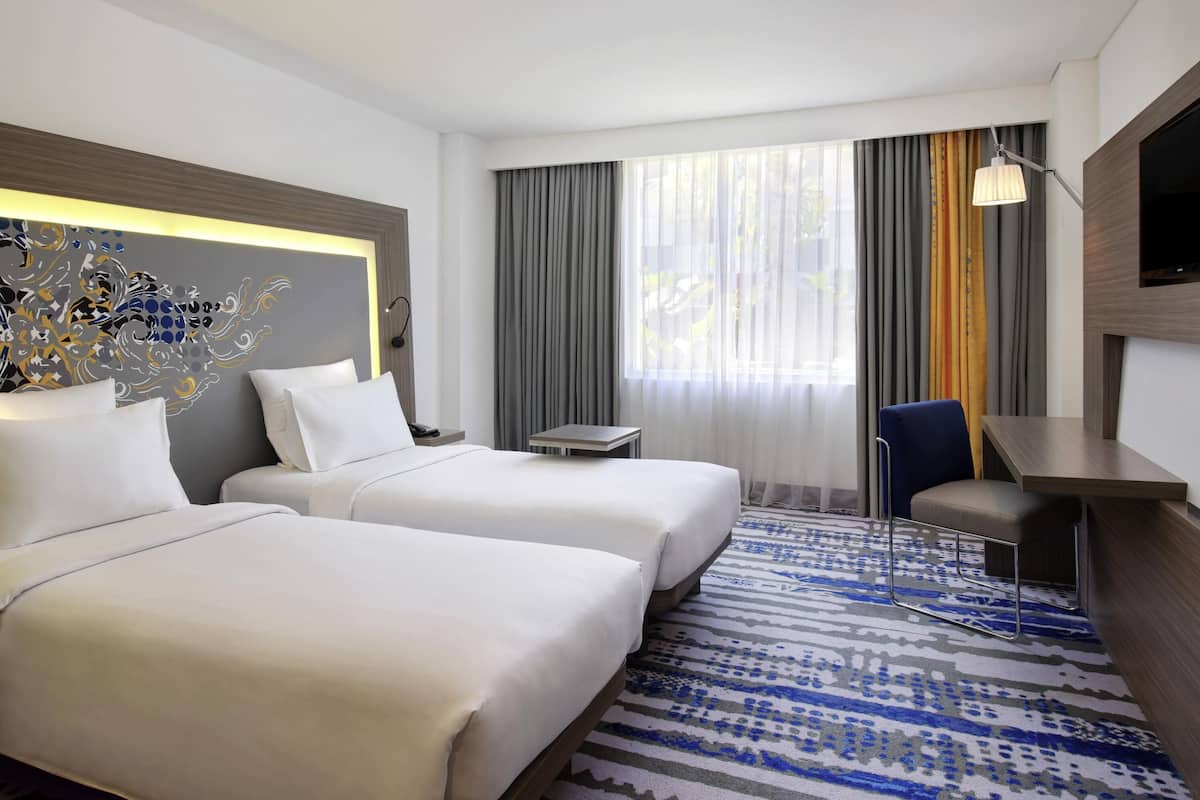 Habitación de hotel con dos camas, un escritorio, una silla, un televisor y un ventanal grande con cortinas.
