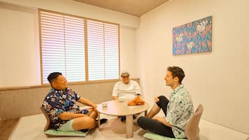 Três pessoas sentadas em esteiras de tatame ao redor de uma mesa redonda em um quarto com uma janela grande e um quadro pendurado na parede.