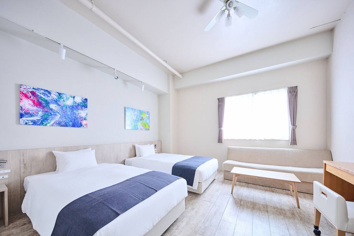 Ein Hotelzimmer mit zwei Betten, einem Schreibtisch, einer Bank und einem Deckenventilator.