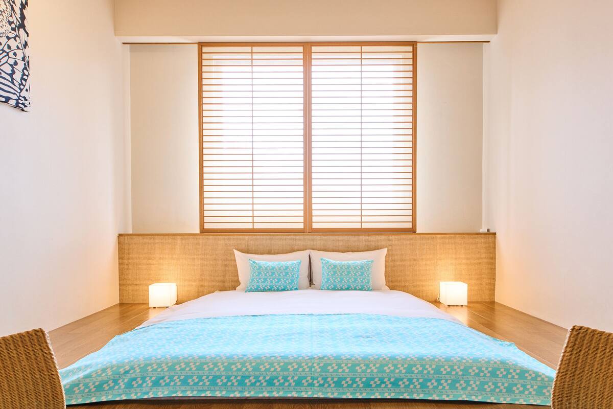 Ein Hotelzimmer mit einem großen Bett, zwei Nachttischlampen, einem Fenster mit Jalousien und einem gewebten Kopfteil.