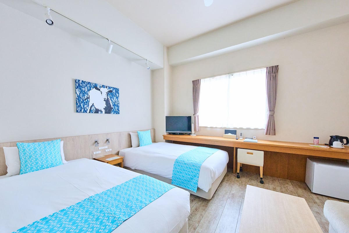 Ein Hotelzimmer mit zwei Betten, einem Schreibtisch mit Computer, einem Fernseher und einem Fenster mit Vorhängen.