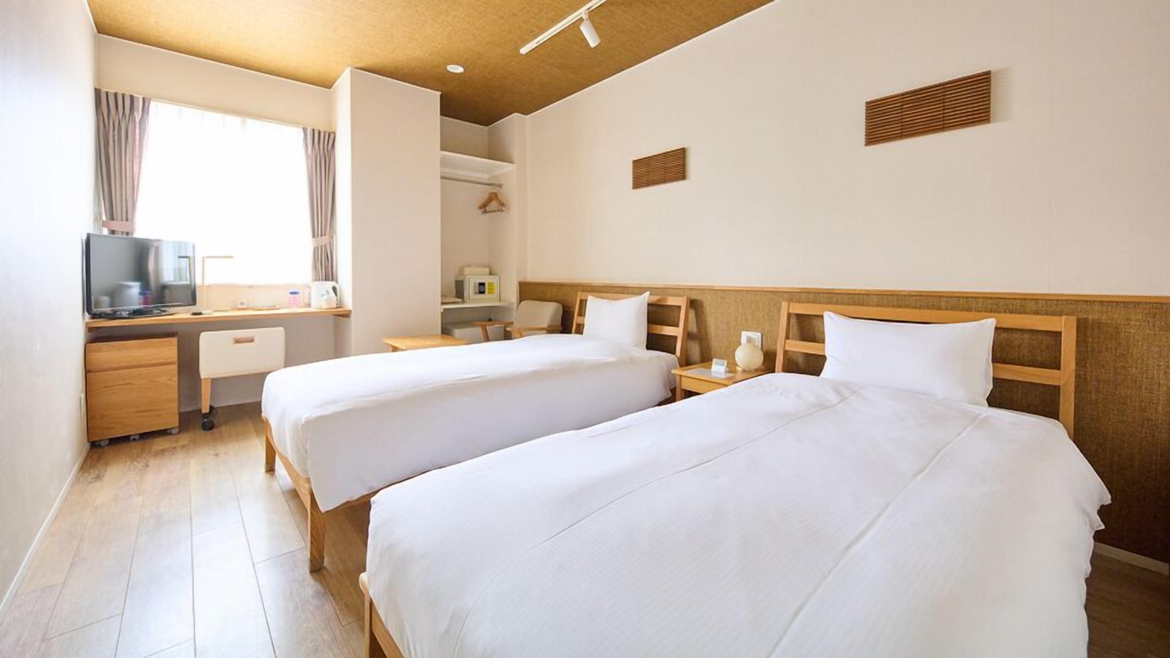 Quarto de hotel com duas camas, uma escrivaninha com televisão, uma janela com cortinas e uma kitchenette pequena.