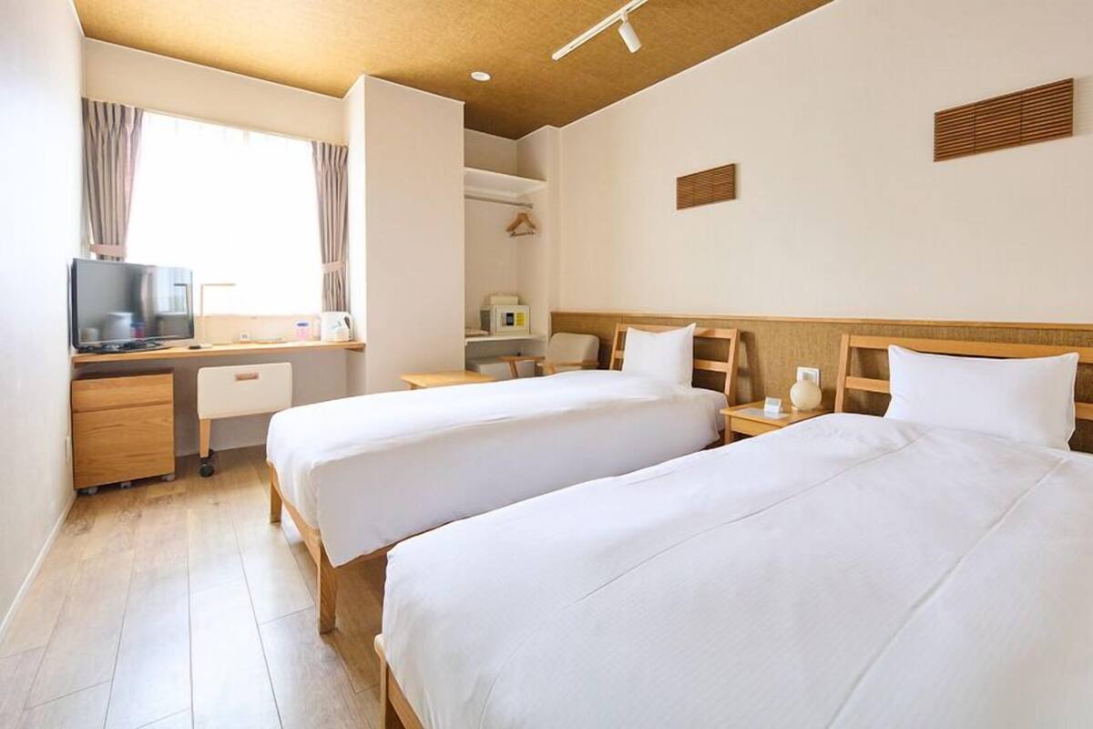 Ein Hotelzimmer mit zwei Betten, einem Schreibtisch mit Fernseher, einem Fenster mit Vorhängen und einer kleinen Küchenzeile.