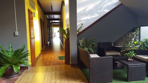Um corredor com piso de madeira, paredes amarelas e plantas em vasos. Há uma área de estar com móveis de vime e vista do céu.